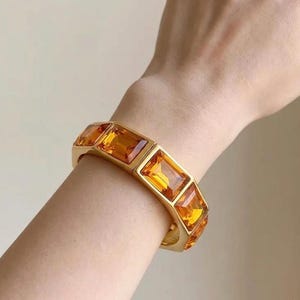 May include: A gold-toned bracelet featuring a series of rectangular, faceted amber-colored glass stones set in a linked metal frame. The bracelet wraps around the wrist, showcasing the vibrant orange hue and reflective quality of the stones.