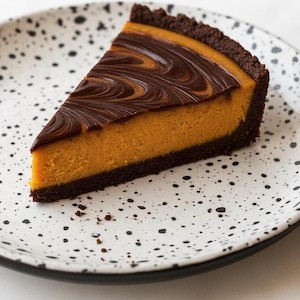 Puede incluir: Una porción de tarta de queso de calabaza con una cobertura de remolino de chocolate y una corteza de chocolate negro. La tarta de queso tiene un relleno naranja y está sobre un plato blanco con motas negras.