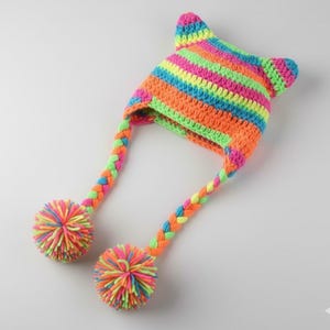 Puede incluir: Un gorro de ganchillo hecho a mano y vibrante con puntas en forma de orejas y lazos trenzados que terminan en pompones. El gorro presenta rayas horizontales en colores neón: rosa, naranja, amarillo, verde y azul. Los pompones y trenzas incorporan la misma paleta de colores brillantes.