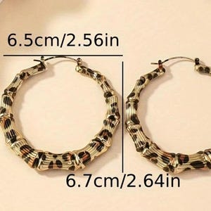 May include: A pair of large gold hoop earrings with a leopard print design. The earrings measure 6.5 cm/2.56 inches in diameter and 6.7 cm/2.64 inches in diameter.