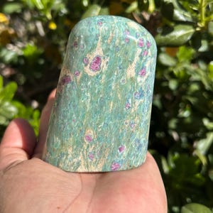 May include: A polished, rounded stone with a teal and beige base color, speckled with vibrant pink and blue inclusions. The stone is held in a hand, with a blurred green foliage background. The stone's shape is smooth and organic, suggesting a decorative or collectible item.