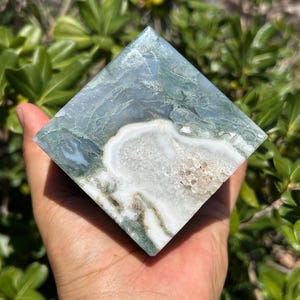 May include: A square-shaped, polished stone with a unique pattern of blue-green and white swirls. The stone is held in a hand, showcasing its natural beauty and intricate design. The stone's edges are sharp and well-defined, highlighting its geometric form.