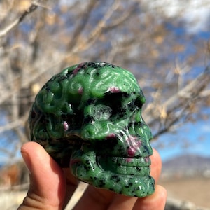 May include: A carved skull made of green and red gemstone. The skull has detailed features, including eye sockets, a nose cavity, and teeth. The gemstone has a polished surface and a natural, organic appearance.