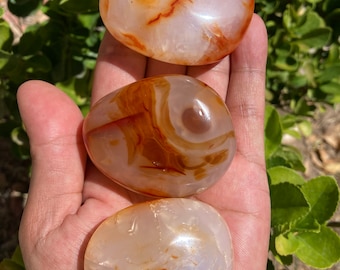 May include: Three carnelian heart-shaped stones with white and orange banding. The stones are smooth and polished.