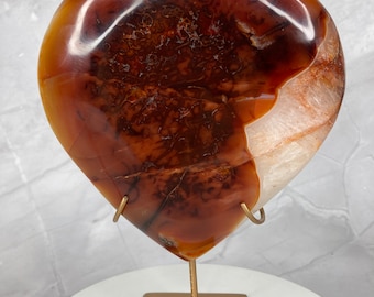 May include: A heart-shaped carnelian stone on a gold metal stand. The carnelian is a deep orange colour with white and brown markings.