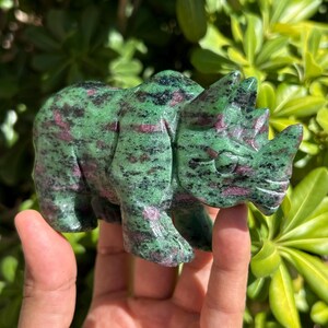 May include: A hand-carved rhinoceros figurine crafted from zoisite stone, featuring a mottled pattern of deep forest green, black, and vibrant magenta-pink inclusions throughout its polished, smooth, and solid mineral body structure.