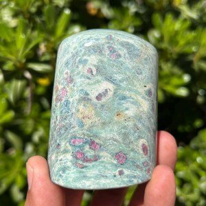 May include: A cylindrical stone with a mottled pattern of teal, light green, and pinkish-red spots. The stone is held in a hand against a backdrop of green foliage. The stone's surface has a slightly polished appearance.