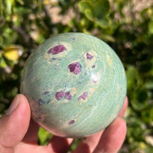 May include: A green sphere with red and pink inclusions. The polished stone is held in a hand, with a blurred green background. The sphere is a natural stone, possibly a decorative item or a collectible.