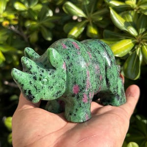May include: A carved stone rhinoceros figurine featuring a vibrant green base color with prominent black speckles and scattered patches of deep magenta. The sculpture displays a smooth, polished texture with detailed facial features and two horns.