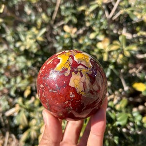 May include: A polished gemstone sphere with a mix of red, yellow, and white colors. The sphere is held in a hand, with a blurred green background of foliage. The stone has a marbled appearance.