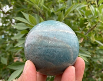 May include: A smooth, green and white striped sphere made of polished stone. The sphere is being held in a hand.