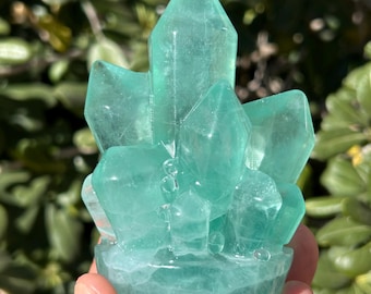 May include: A translucent, light green crystal cluster sculpture. The crystal formation has multiple pointed facets and sits on a rounded base. The sculpture is held in a hand against a blurred green foliage background.