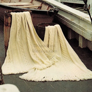 May include: Two cream-colored crocheted blankets with fringe, draped over wooden barrels, against a backdrop of a wooden boat and a weathered dock.