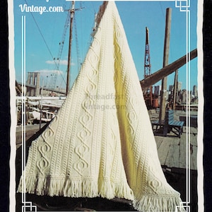 May include: Cream-colored knitted blanket with a cable knit design and fringe. The blanket is draped over a wooden structure with a cityscape and blue sky in the background. The text "Threadfast Vintage.com" is visible.