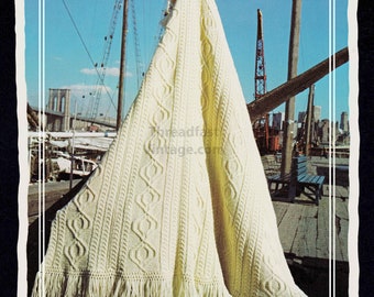 May include: Cream-coloured knitted blanket with a cable knit design and fringe. The blanket is draped over a wooden structure with a cityscape and blue sky in the background. The text "Threadfast Vintage.com" is visible.