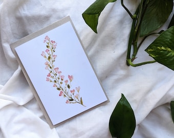 May include: A rectangular greeting card featuring a watercolor illustration of a delicate, curving stem with small pink blossoms and green leaves. The card is centered on a light gray backing, showcasing a clean, white surface for the artwork.
