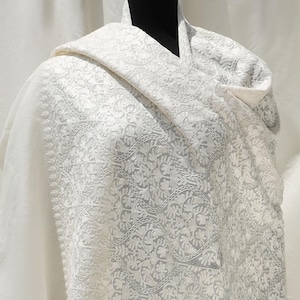 May include: A white shawl with a delicate floral pattern in a lighter shade of white. The shawl is draped over a mannequin and has a fringed edge.