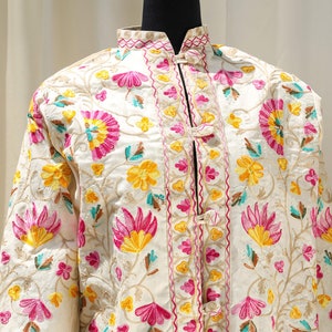 May include: A white silk jacket with a mandarin collar and floral embroidery. The jacket has a button closure and is decorated with pink, yellow, and green flowers.