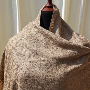 May include: A beige shawl with an intricate embossed floral pattern. The shawl is draped over a mannequin, showcasing the detailed design and texture of the fabric. The overall aesthetic is elegant and sophisticated.