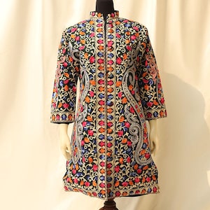 Pode incluir: Uma jaqueta preta de manga comprida com gola mandarim e bordados florais intrincados em tons de azul, rosa, laranja e dourado sobre um fundo verde escuro.