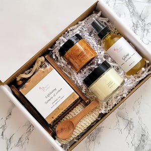 May include: A gift box containing skincare products. Includes a bar of exfoliating turmeric and lemon soap, a jar of scrub, a bottle of Glow spray, a jar of turmeric body butter, a wooden spoon, and a loofah. The box is filled with white paper and has a marble background.