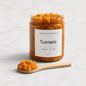 Puede incluir: Un frasco de plástico transparente lleno de exfoliante de azúcar y cúrcuma naranja, con una etiqueta blanca que dice Water For Roses, Turmeric, sugar scrub, 8oz. Una pequeña cuchara de madera contiene una porción del exfoliante.