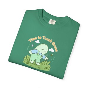 May include: Green t-shirt featuring the text "Time to Touch Grass" and a cartoon character in dungarees reaching for grass. The design includes butterflies and clouds, creating a whimsical scene.