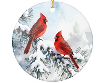May include: A round ceramic ornament featuring two red cardinal birds perched on a snow-covered pine branch. The birds have black beaks and crests. The background is a watercolor winter scene with a gold string for hanging.
