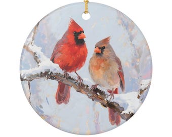 May include: Round ceramic ornament featuring a painting of two cardinals perched on a snow-covered branch. The male cardinal is bright red with a black mask, while the female is a muted brown. A gold string is attached for hanging.