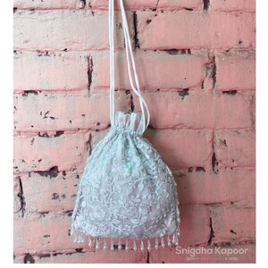 May include: A white drawstring bag with a delicate floral pattern and pearl accents. The bag is hanging from a white cord against a pink brick wall.