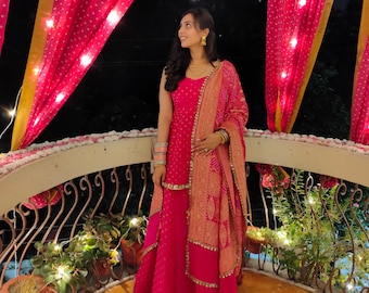 May include: A bright pink sleeveless kurta and matching skirt set featuring a subtle dotted pattern, paired with a long, ornate peach and gold embroidered dupatta with a decorative border, gold jewelry, and stacked bangles on both wrists.