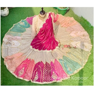 Op de afbeelding: Een levendige, veelkleurige traditionele Indiase outfit. De set bestaat uit een beige top met gouden borduursels, een felroze dupatta en een rok met panelen in roze, groen en perzik. De rok heeft ingewikkelde gouden details.