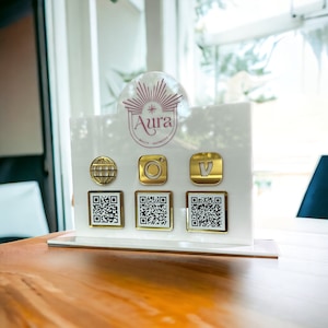 May include: White acrylic sign with the logo "Aura Beauty - Aesthetics" and a gold star. The sign has four gold icons: a globe, an Instagram camera, a Vimeo logo, and a QR code. There are three QR codes on the sign.