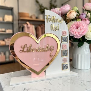 May include: A pink heart-shaped sign with gold trim and the word "Libertad" in gold script. The sign also features the words "Follow Us!" and social media QR codes. A bouquet of pink and white flowers sits nearby.
