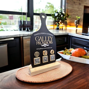 May include: A black cutting board sign with gold lettering that reads "CHARCUTERIE & CATERING GALLEY PROVISIONS www.galleyprovisionsmaine.com". The sign has four gold icons with QR codes below them. The icons are a globe, an Instagram logo, a Facebook logo, and a QR code.