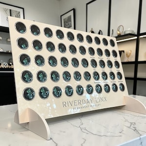 May include: A display board for permanent jewelry featuring a grid of teal gemstones, each set in a silver frame. The board is cream-colored with the text "RIVERDAY LINX PERMANENT JEWELRY" and the months of the year. The display is supported by a white stand.