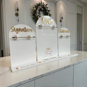 May include: Three white acrylic display stands with gold lettering that reads "Signature", "Icon", and "Statement". The stands have a gold bar for displaying jewelry. The brand name "Links and Chains" is also visible.