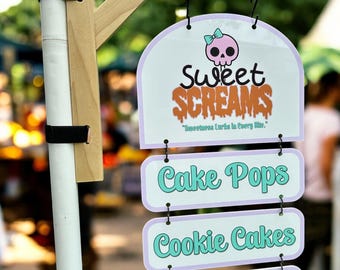 May include: A sign for "Sweet Screams" featuring a skull with a bow, and the text "Cake Pops", "Cookie Cakes", and "Ditty Cakes". The sign is white with a purple border and is hanging from a wooden structure.