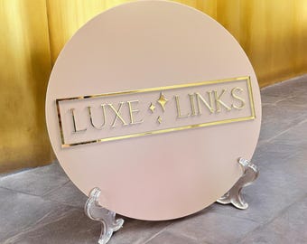 Può includere: Un cartello rotondo di colore rosa chiaro con un bordo dorato e il testo "LUXE LINKS" in lettere dorate. Il cartello è su un supporto in acrilico trasparente.