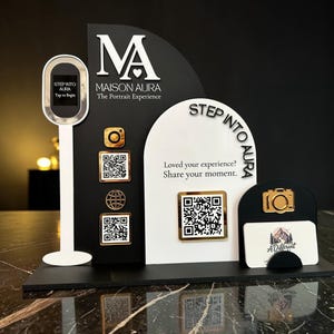 May include: A black and white display stand with the text "MAISON AURA The Portrait Experience" and "STEP INTO AURA". It features a photo booth, QR codes, and a business card holder with a camera icon.