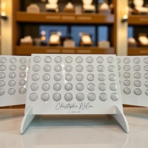 May include: White display stands with rows of clear, circular objects. The stands have a minimalist design with a clean, modern aesthetic. The name "Christopher Nolan" is written on the front.