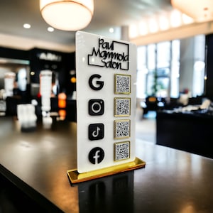 May include: A white sign with the text "Paul Mammola Salon" and five QR codes for different social media platforms: Google, Instagram, TikTok, Facebook. The sign is on a black surface.
