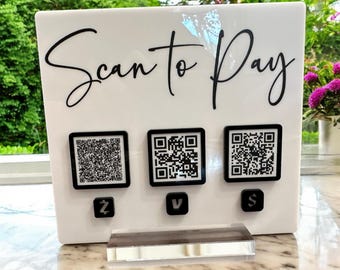 Può includere: Un cartello bianco con il testo "Scan to Pay" in scrittura corsiva nera. Tre codici QR in bianco e nero sono visualizzati sul cartello, ognuno con un'icona di metodo di pagamento diversa sotto: Z, V e $.