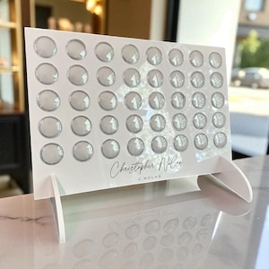 May include: A white acrylic display with a grid of clear, circular elements. The display is supported by two white stands. The name "Christopher Nolan" is written in elegant script. The display is on a reflective surface.