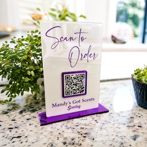 May include: A white sign with purple text and a purple border. The sign says "Scan to Order" and has a QR code. Below the QR code is the text "Mandy's Got Scents Scentsy".