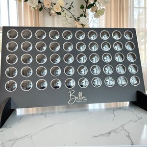 May include: A dark gray Connect Four game board with clear, reflective game pieces. The board has a black base and the word "Bella" in cursive script. A classic game for family fun.