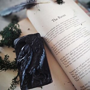 Puede incluir: Un objeto decorativo negro rectangular con tem&aacute;tica de cuervo, con intrincadas tallas, apoyado en un libro abierto. Las p&aacute;ginas del libro muestran el poema "El Cuervo" de Edgar Allan Poe. Musgo verde y una pluma a&ntilde;aden a la est&eacute;tica g&oacute;tica.