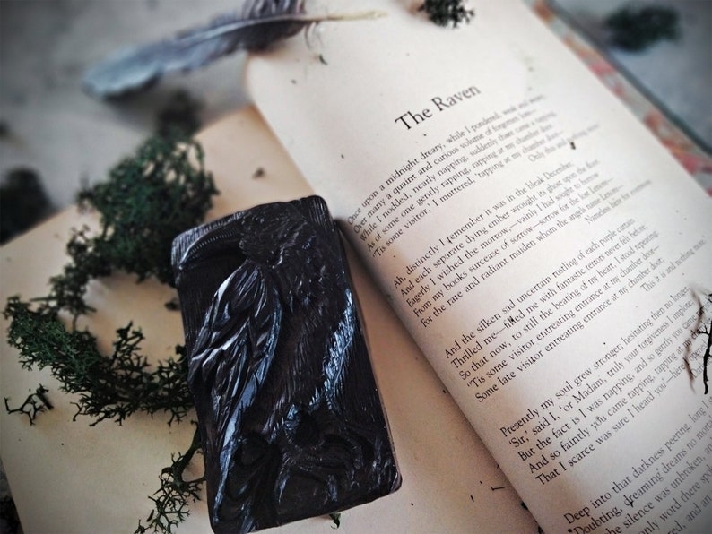 Puede incluir: Un objeto decorativo negro rectangular con tem&aacute;tica de cuervo, con intrincadas tallas, apoyado en un libro abierto. Las p&aacute;ginas del libro muestran el poema "El Cuervo" de Edgar Allan Poe. Musgo verde y una pluma a&ntilde;aden a la est&eacute;tica g&oacute;tica.
