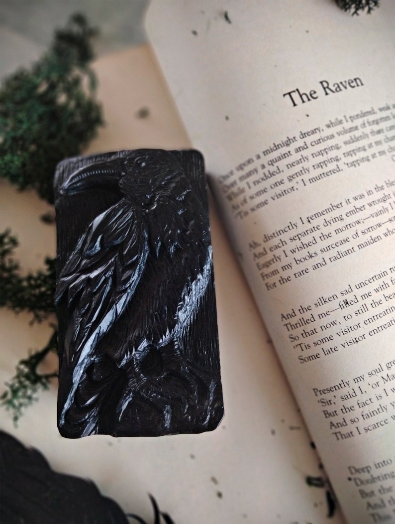 Puede incluir: Un objeto decorativo o jab&oacute;n rectangular negro con un dise&ntilde;o de cuervo tallado. El cuervo est&aacute; representado con plumas detalladas y un pico prominente. El objeto se coloca sobre un libro abierto con el t&iacute;tulo "The Raven".