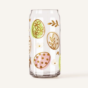 Op de afbeelding: Een transparante glazen kan met een gouden ontwerp van paaseieren en bloemmotieven. De eieren zijn versierd met stippen, bloemen en bladeren.